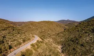 Vue aérienne d'une route sinueuse traversant des collines verdoyantes, menant vers le col du Testanier. Deux cyclistes s'engagent sur la pente.