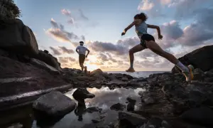 Deux coureurs s'entraînent sur un sentier rocailleux à Tenerife au coucher du soleil, l'océan en arrière-plan.