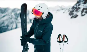 Femme en tenue de ski, tenant ses skis, portant un casque de ski avec visière intégrée.