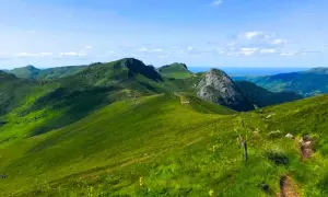Paysage de montagnes verdoyantes sous un ciel bleu, idéal pour les plus belles randonnées dans le Cantal. Un sentier serpente sur la colline.