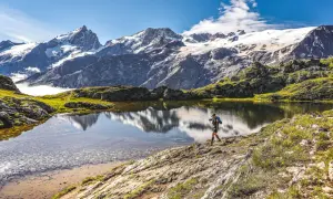 Un randonneur traverse le plateau d'Emparis, un lac alpin reflétant des montagnes enneigées sous un ciel bleu.