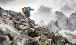 Un randonneur avec un grand sac à dos escalade un sentier rocheux, les pics des Dolomites se dressent dans le brouillard.
