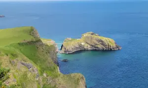 Falaises verdoyantes de la Causeway Coast, avec une île rocheuse reliée par un pont suspendu. L'eau bleue claire invite à la découverte.