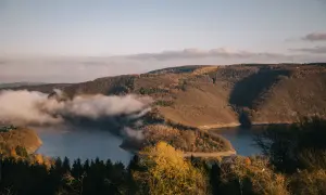 Vue panoramique d'un lac sinueux entouré de collines boisées aux couleurs d'automne, avec des nuages bas flottant sur l'eau. Un paysage typique du parc national de l'Eifel.