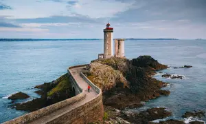 Une personne en manteau rouge marche sur la route des phares, un chemin sinueux menant à un phare sur une île rocheuse.