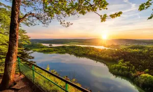 Vue panoramique depuis le belvédère Pic de l'Aigle au coucher du soleil, avec un lac reflétant le ciel et des collines verdoyantes.
