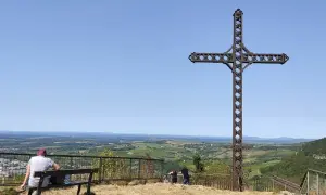 La croix du Dan domine un paysage verdoyant. Deux personnes profitent de la vue depuis un banc.
