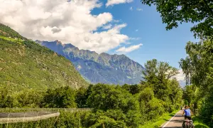 Cycliste sur la Via Claudia Augusta, traversant une vallée verdoyante bordée de montagnes majestueuses.