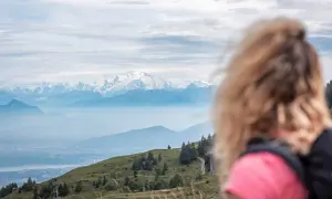Vue panoramique sur les Alpes enneigées, avec une femme blonde en premier plan. Un sentier serpente sur la colline verdoyante, invitant au trek dans le Jura.