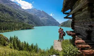 Un couple avec un bébé admirent le lac d'Aoste, entouré de montagnes verdoyantes et d'une cabane en bois rustique.