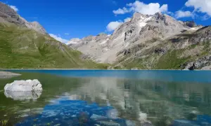 Le lac de la Sassière, aux eaux turquoise, reflète les montagnes majestueuses et le ciel bleu. Un rocher émerge, témoin de la quiétude du paysage.