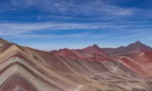 La montagne Vinicunca, aux couleurs vives, s'étend sous un ciel bleu parsemé de nuages. Un sentier serpente à travers ses flancs striés.
