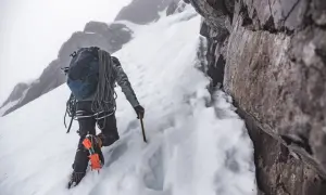 Grimpeur en crampons et piolet, progressant sur une pente enneigée. Un entraînement en altitude exigeant, au cœur d'un paysage rocheux et brumeux.
