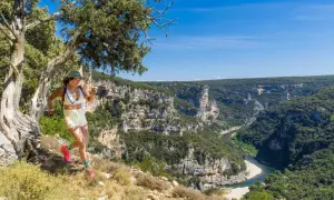 Une femme court sur un sentier escarpé avec une vue spectaculaire sur le **trail gorge de l'Ardèche**. Le soleil brille sur les falaises et la rivière.