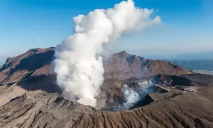 Fumée s'échappant du cratère du volcan Aso, sous un ciel bleu clair avec la lune visible.