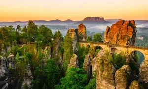 Un pont de pierre traverse des formations rocheuses spectaculaires, rappelant la Suisse bohémienne. La nature y est luxuriante, baignée par la lumière dorée du crépuscule.