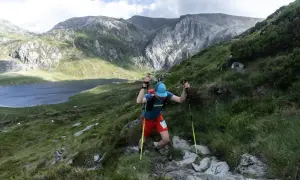 Un coureur grimpe une pente rocheuse, bâtons en main, lors de la kat100 by utmb. Un lac et des montagnes majestueuses en arrière-plan.