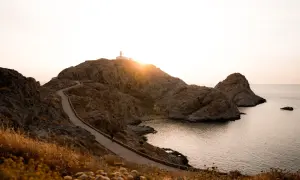 Chemin sinueux menant à un phare sur une colline rocheuse, une porte de pierre naturelle s'ouvrant sur la mer au coucher du soleil.