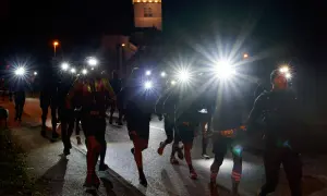 Courseurs de trail de nuit, lampes frontales allumées, traversant un village sombre. Un chien les accompagne.