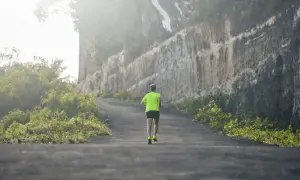 Un coureur en tenue fluo grimpe une route sinueuse, affrontant un dénivelé positif de 100 m, bordée de végétation luxuriante et d'une falaise imposante.