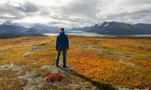 Une randonneuse admire un paysage automnal aux couleurs vives, parfait pour randonner en novembre en France. Montagnes enneigées et lac en arrière-plan.