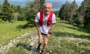 Un coureur en tenue de trail, sac à dos rouge, sur un sentier escarpé près d'Oyonnax, profitant d'un paysage verdoyant et vallonné.