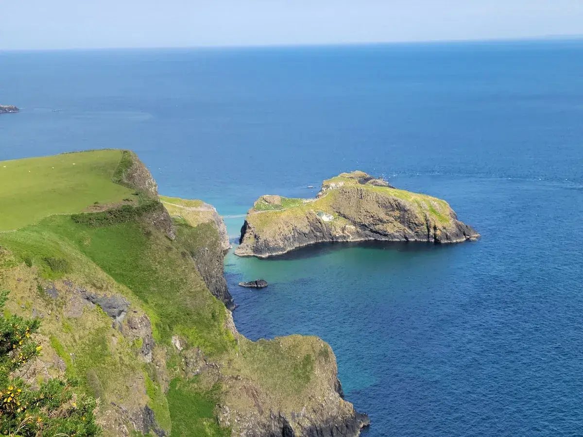 Falaises verdoyantes de la Causeway Coast, avec une île rocheuse reliée par un pont suspendu. L'eau bleue claire invite à la découverte.