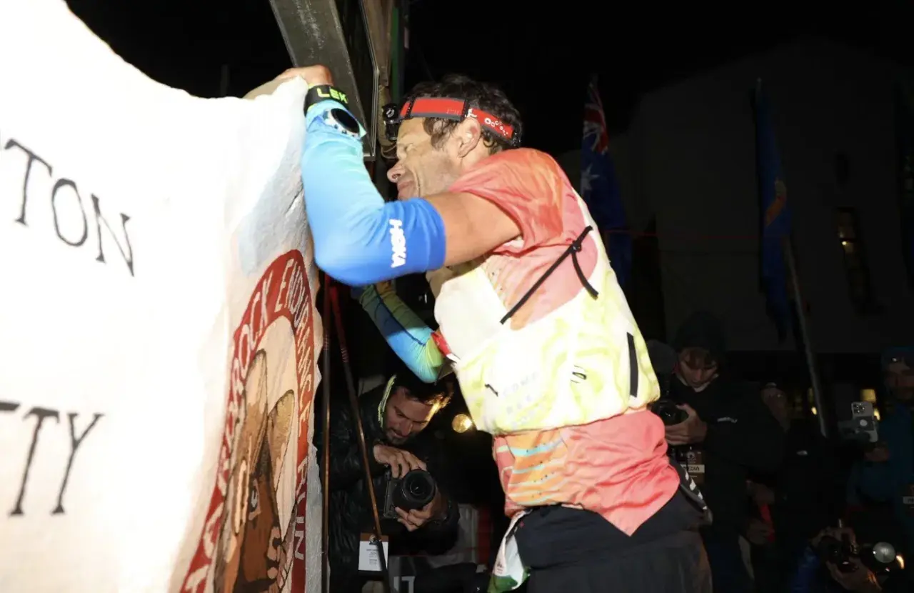 Un coureur, épuisé mais victorieux, est acclamé lors du record Hardrock 100. Des drapeaux flottent derrière lui.