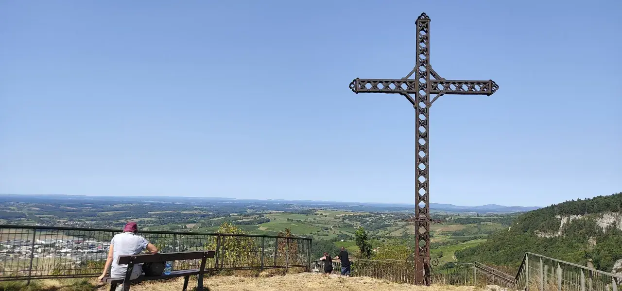 La croix du Dan domine un paysage verdoyant. Deux personnes profitent de la vue depuis un banc.