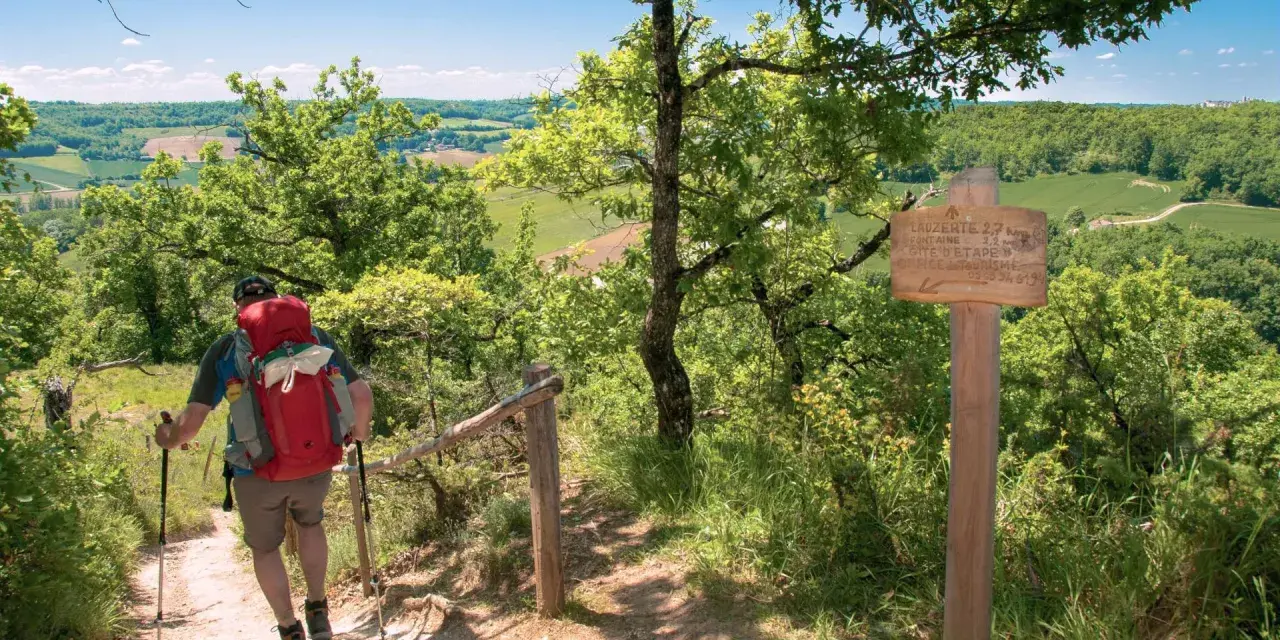 Randonnées Tarn et Garonne : un marcheur avec un sac à dos rouge monte un sentier boisé, dominant un paysage verdoyant. Panneau indicateur de Lauzerte.