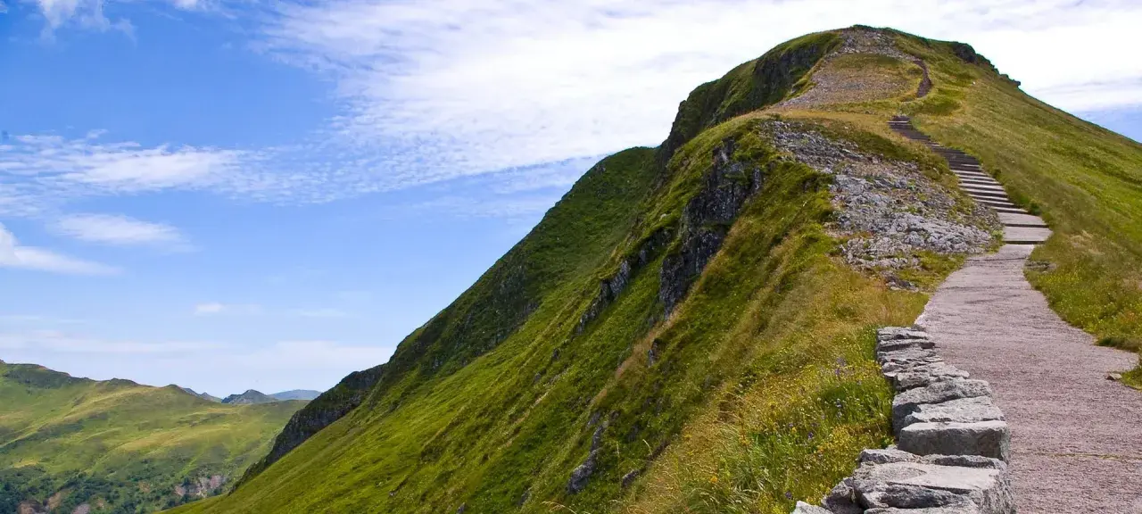 Sentier rocailleux menant au sommet d'une montagne verdoyante en gr France, sous un ciel bleu parsemé de nuages.