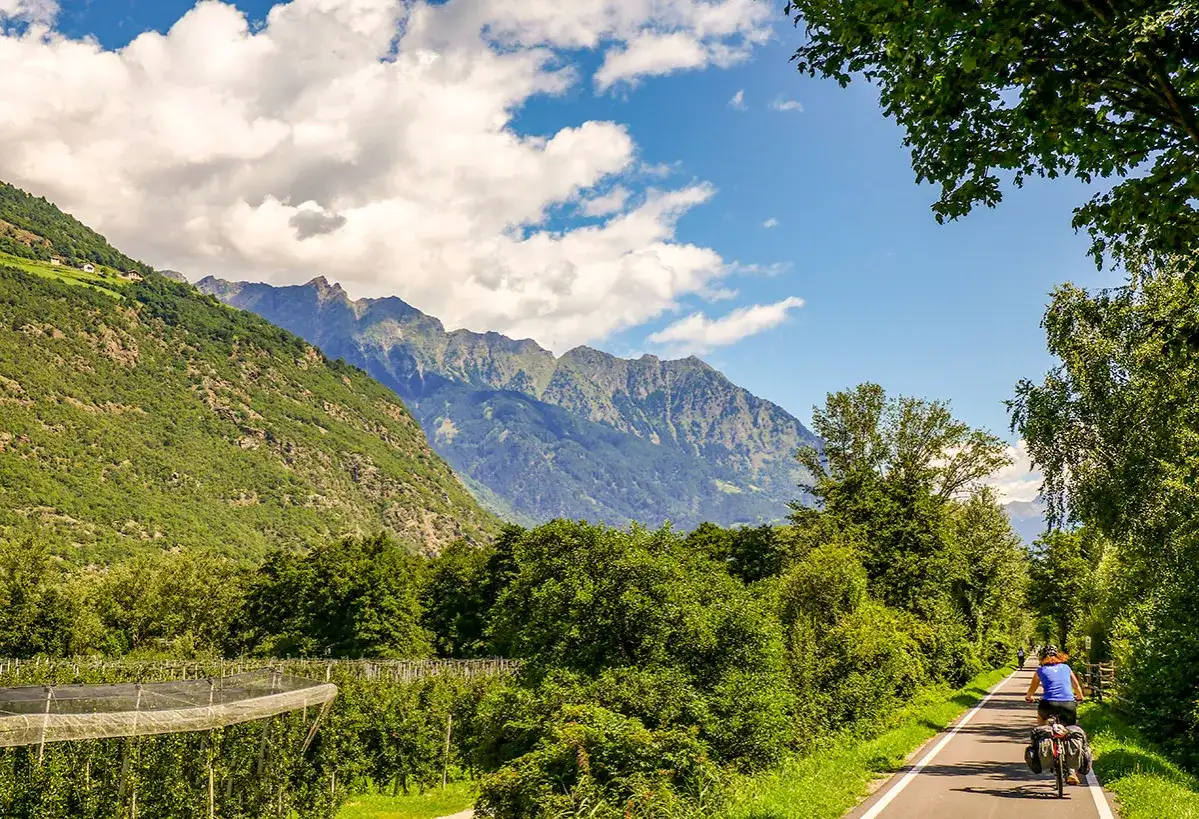 Cycliste sur la Via Claudia Augusta, traversant une vallée verdoyante bordée de montagnes majestueuses.