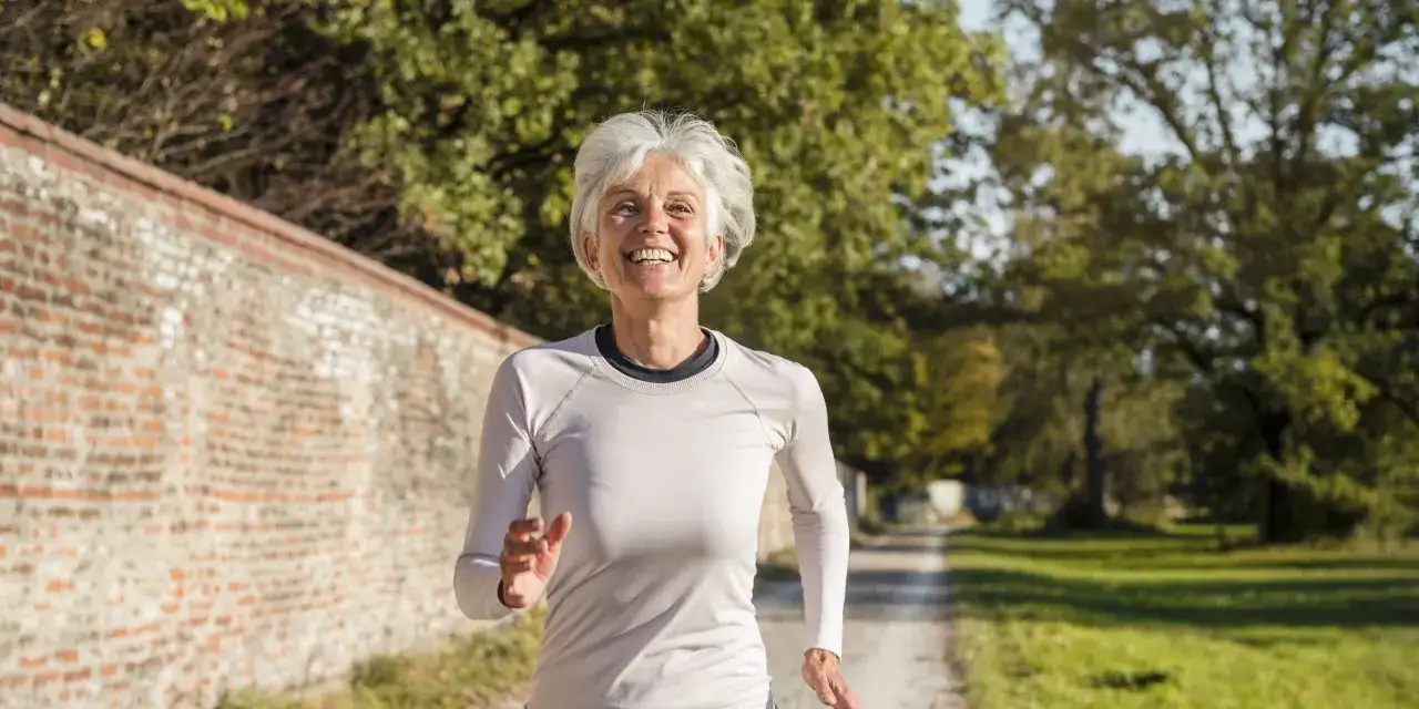 Femme souriante de 60 ans courant sur un chemin, suivant un plan d'entraînement course à pied.