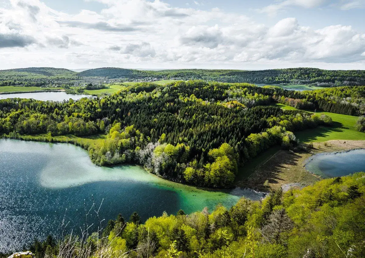 Vue aérienne d'un lac aux eaux turquoise, entouré de forêts verdoyantes et de collines. Un paysage idéal pour un trek dans le Jura.