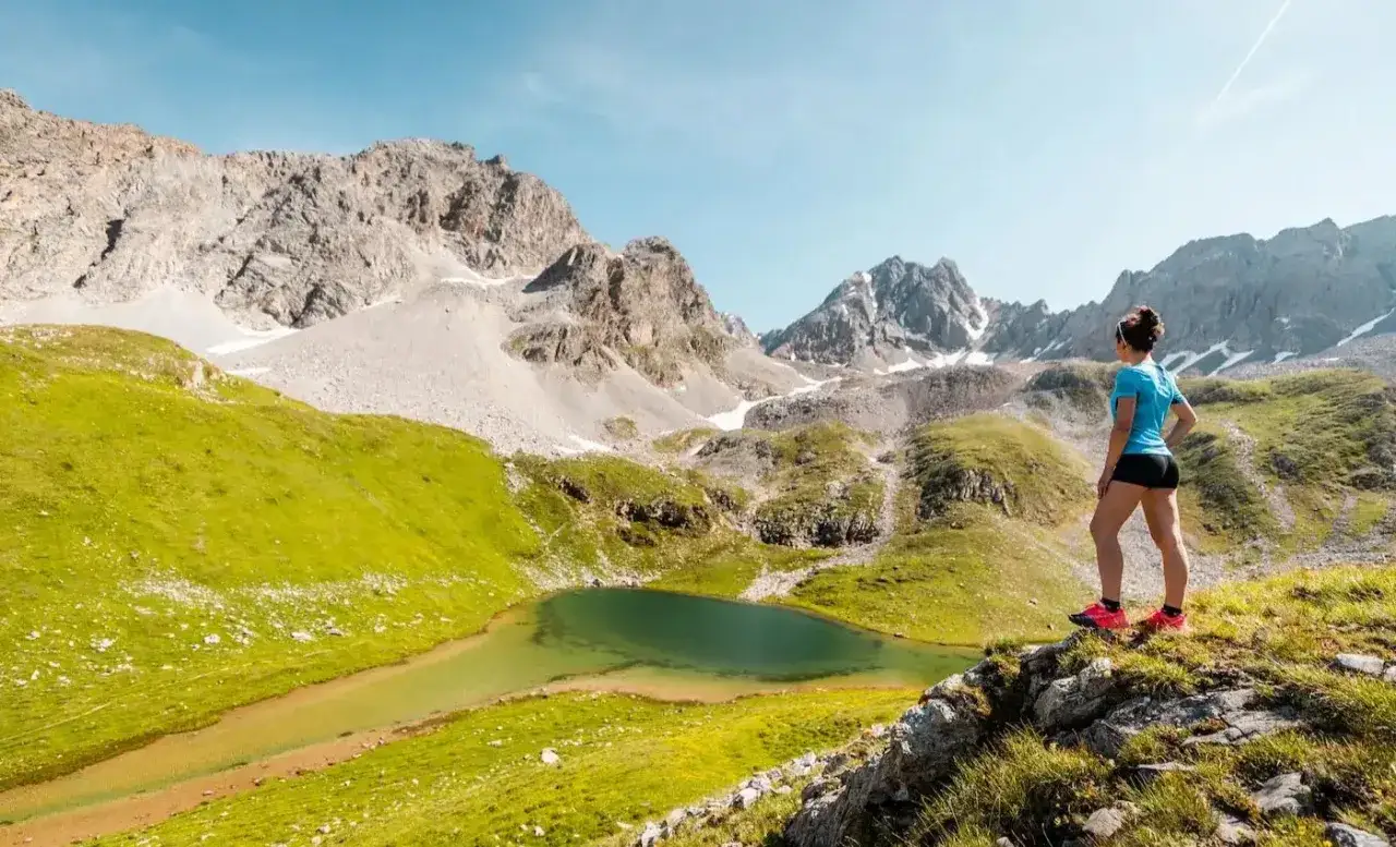 Une femme admire un lac de montagne lors d'une randonn&eacute;e &agrave; Courchevel. Le paysage est spectaculaire avec des montagnes rocheuses et des prairies verdoyantes.