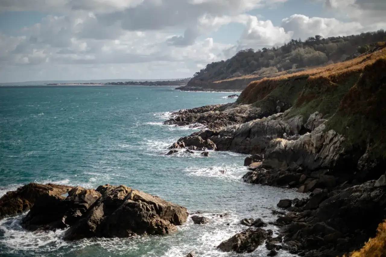 Côte rocheuse battue par les vagues de la Manche, avec une colline verdoyante et des nuages dans le ciel.