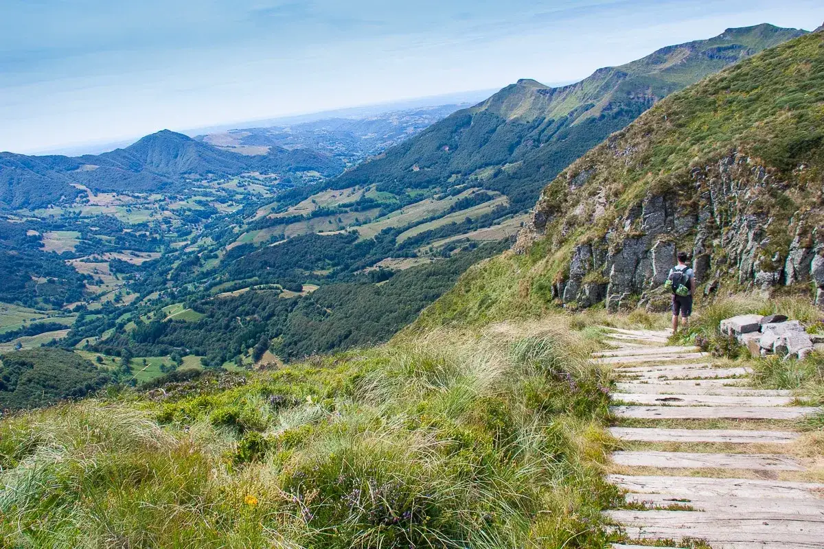 Un randonneur sur un sentier en bois, profitant des plus belles randonn&eacute;es dans le Cantal. Paysage de montagnes verdoyantes et de vall&eacute;es.