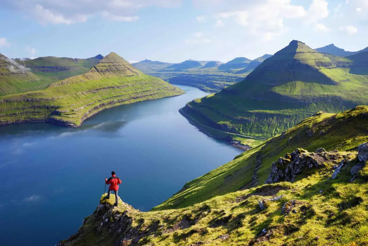 Un randonneur admire un paysage spectaculaire des &icirc;les F&eacute;ro&eacute;, avec des montagnes verdoyantes et un fjord bleu.