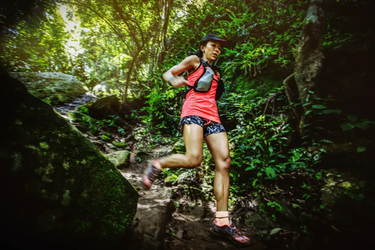 Une coureuse agile d&eacute;vale un sentier forestier, concentr&eacute;e sur sa descente trail. La v&eacute;g&eacute;tation luxuriante l'entoure.