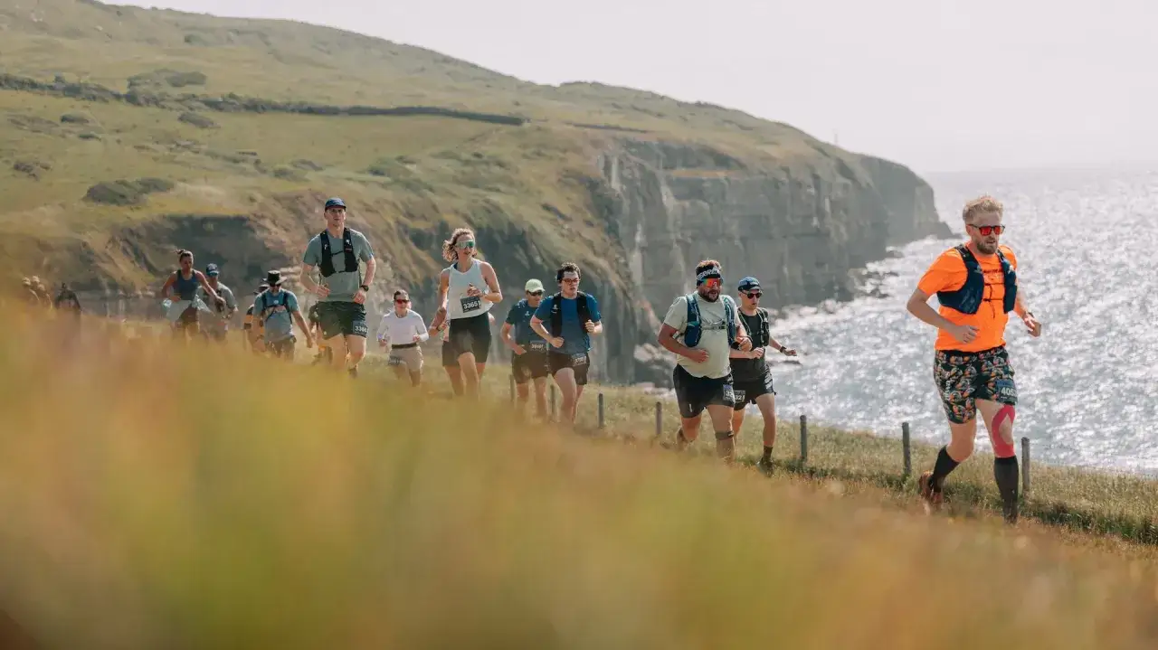 Des coureurs s'élancent sur un sentier côtier, visant un record de 10 km. Le soleil brille sur la mer scintillante et les falaises verdoyantes.