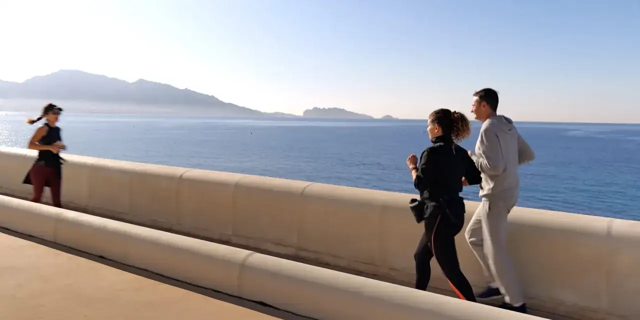 Trois personnes courent le long d'une promenade avec la mer et les montagnes en arrière-plan. Un endroit idéal pour savoir où courir à Marseille.
