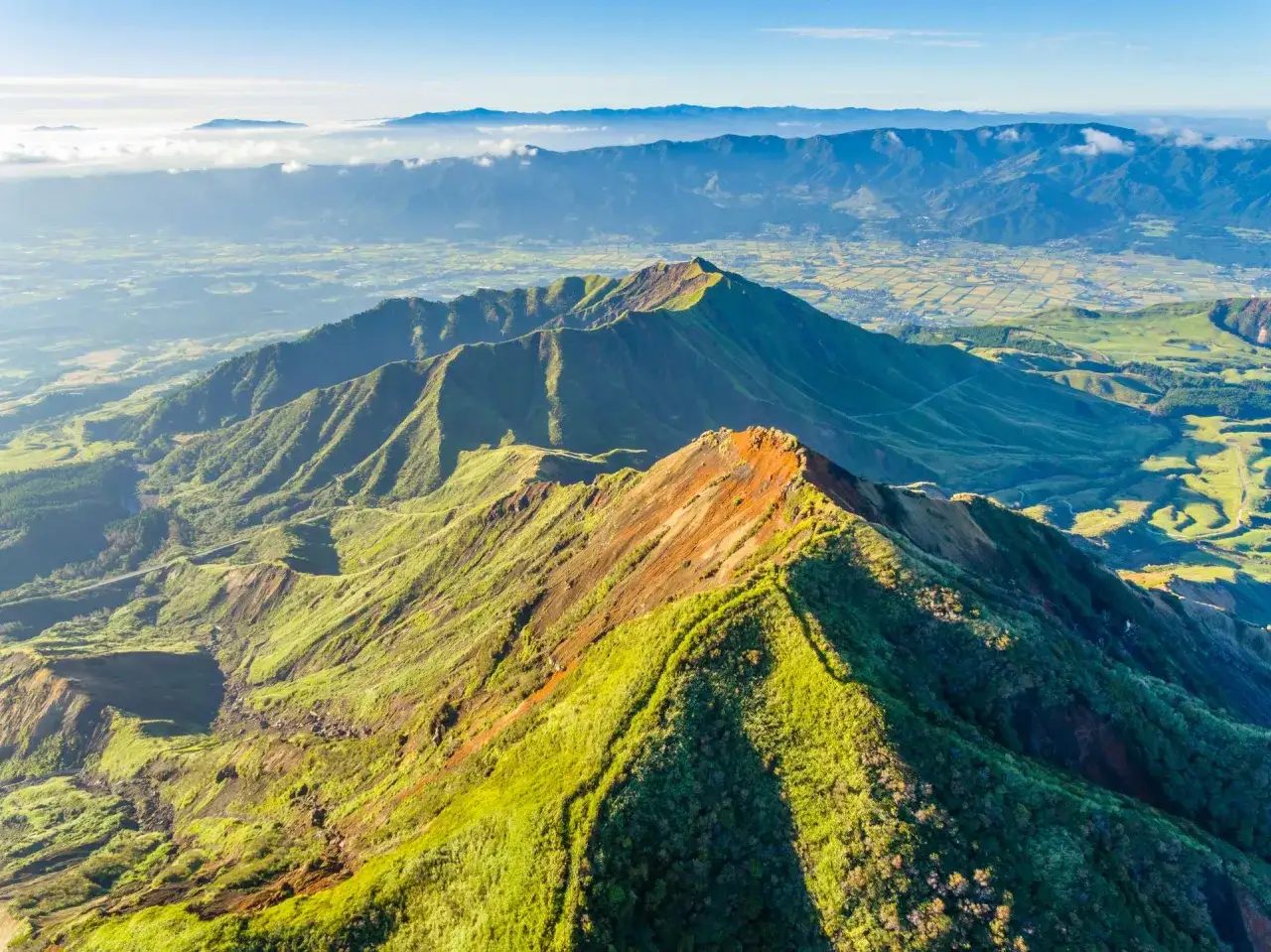 Vue aérienne d'un aso volcan verdoyant, ses crêtes déchiquetées baignées de soleil, dominant une vallée fertile et des montagnes lointaines.