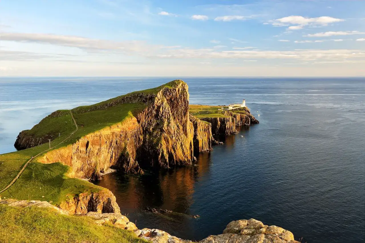 Phare blanc sur une falaise verdoyante de l'île de Skye, baignée par la lumière dorée du soleil couchant. L'océan bleu s'étend à perte de vue.