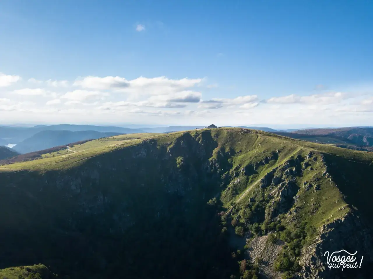 Vue panoramique des Vosges sous un ciel bleu et nuageux. Un sentier serpente sur une crête verdoyante menant à un refuge.