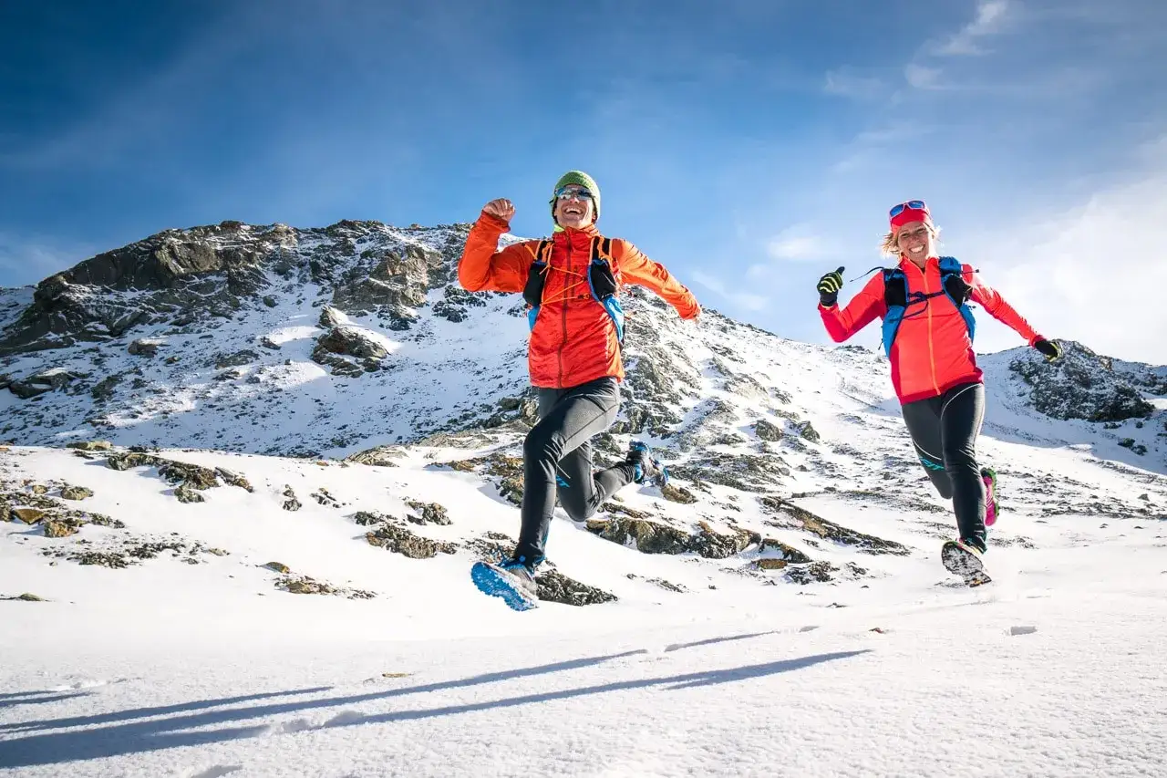 Deux coureurs s'amusent &agrave; courir sous la neige dans un paysage montagneux enneig&eacute;, sous un ciel bleu &eacute;clatant.