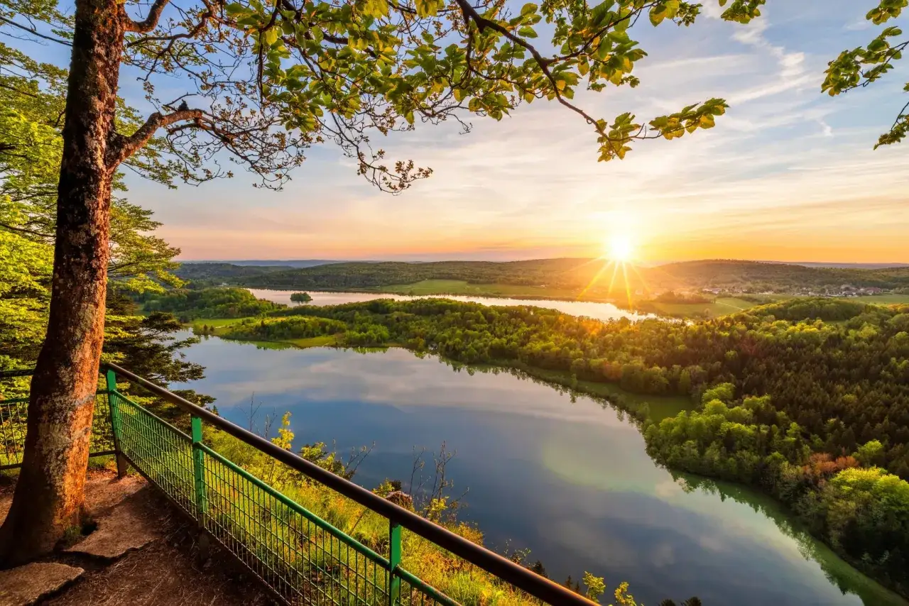Vue panoramique depuis le belvédère Pic de l'Aigle au coucher du soleil, avec un lac reflétant le ciel et une forêt verdoyante.