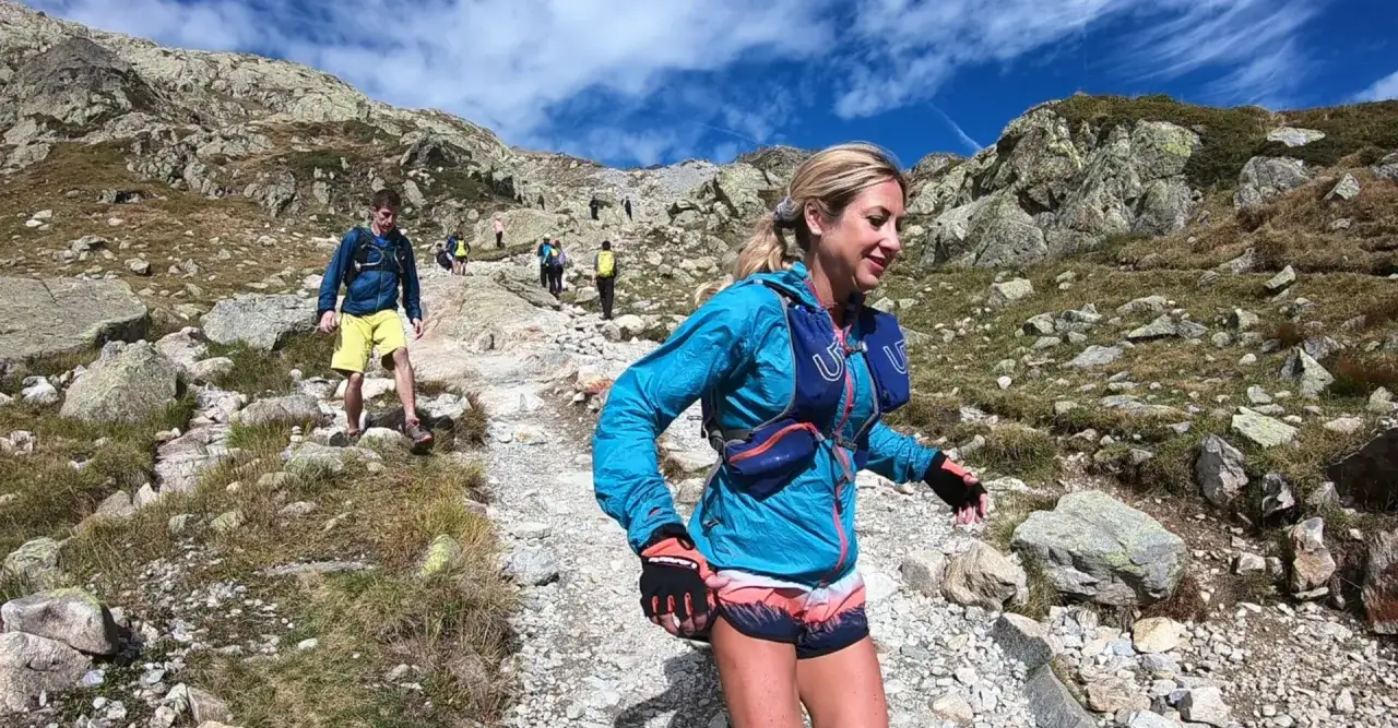 Une femme souriante participe &agrave; un entra&icirc;nement trail en montagne, suivie par d'autres coureurs sur un sentier rocailleux.