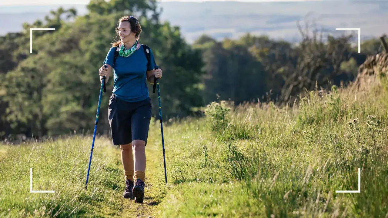 Une femme souriante profite d'une promenade en nature, bâtons à la main. Idéal pour le marche nordique débutant.