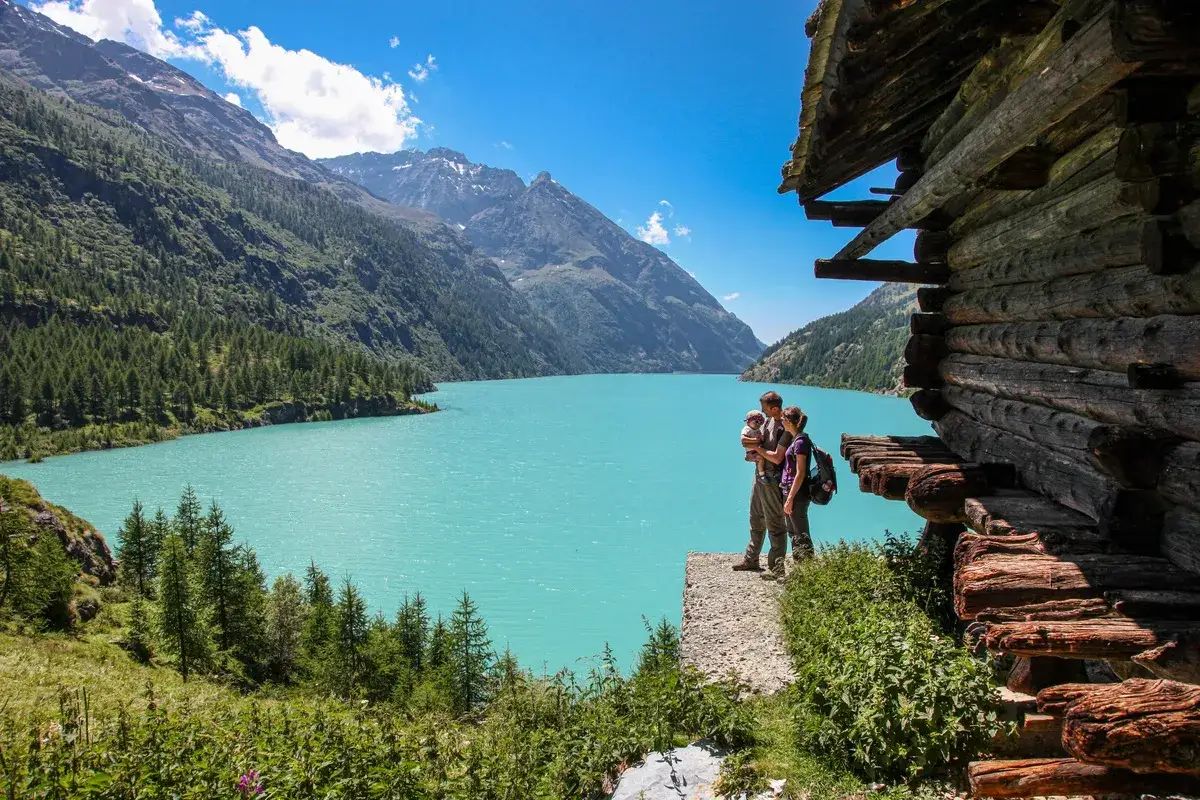 Un couple avec un b&eacute;b&eacute; admirent le lac d'Aoste, entour&eacute; de montagnes verdoyantes et d'une cabane en bois rustique.