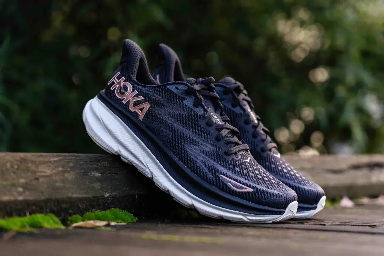 Paire de chaussures de course Hoka Clifton 9 bleu marine avec logo rose sur un fond de verdure. Un avis sur ces chaussures est à venir.
